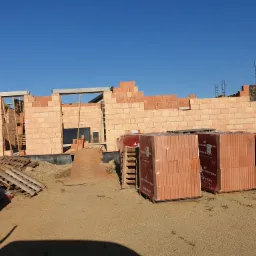 Wznoszenie ścian zewnętrznych z czerwonej cegły Porotherm, widoczne palety z materiałem budowlanym na placu budowy, elementy szalunków i zbrojenia.