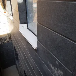 Elewacja budynku z ciemnymi, poziomymi panelami, okno zabezpieczone folią ochronną, widoczne kable i instalacje w tle.