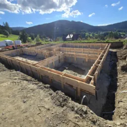 Zalane fundamenty domu jednorodzinnego z drewnianym szalunkiem, widok z góry na tle górskiego krajobrazu w słoneczny dzień. Etap budowy.