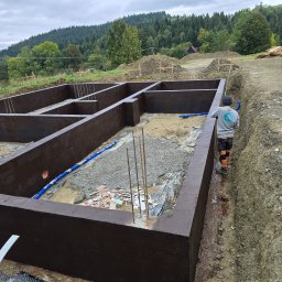 Modernbau Sylwester Bielski - Zabezpieczone, pomalowane fundamenty domu jednorodzinnego na tle górskiego krajobrazu. Widoczny pracownik w odzieży roboczej i niebieski wąż.