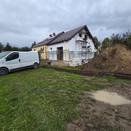 Modernbau Sylwester Bielski - Ocieplanie elewacji domu jednorodzinnego z żółtą dobudówką. Na rusztowaniu pracownicy montują styropian. Obok sterta ziemi i biały bus.