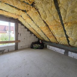 Modernbau Sylwester Bielski - Poddasze ocieplone wełną mineralną w Gdowie. Widoczne bloczki betonowe ścian i okno z widokiem na zewnątrz. Izolacja termiczna zamontowana między krokwiami dachu.