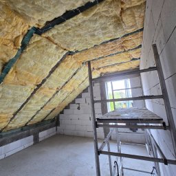 Modernbau Sylwester Bielski - Poddasze w trakcie ocieplania wełną mineralną, widoczne krokwie i sznurki mocujące izolację. Ściany z białych bloczków, drabina aluminiowa w pomieszczeniu.