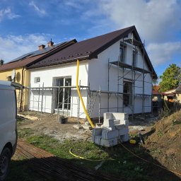 Modernbau Sylwester Bielski - Ocieplony dom w trakcie budowy, z widocznymi rusztowaniami i stertą bloczków. Żółty wąż leży na trawie. Częściowo widoczny biały van.