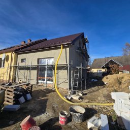 Modernbau Sylwester Bielski - Budynek w trakcie ocieplania elewacji, widoczne rusztowania, węże do zaprawy, materiały budowlane. Częściowo otynkowana ściana i nowy, brązowy dach.