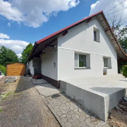 Dom jednorodzinny z białymi ścianami i czerwonym dachem, widok z zewnątrz na podmurówkę i fragment elewacji po remoncie, z widocznym garażem w tle i częściowo ułożonym chodnikiem z płyt.