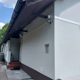 Boczny widok na dom z jasną elewacją, ciemnobrązowym cokołem i drewnianym podbitką dachu, widoczny reflektor zewnętrzny, schody wejściowe i zbiornik na deszczówkę.