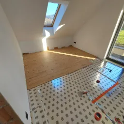 Układanie paneli podłogowych w pokoju na poddaszu z oknem dachowym i dużym oknem balkonowym, widoczne narzędzia i folia izolacyjna pod panelami.