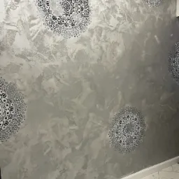 Ściana pokryta dekoracyjną, srebrzystą farbą z naniesionymi, symetrycznymi wzorami mandali w kolorze czarnym, widoczna z perspektywy narożnika pomieszczenia, z fragmentem marmurowej podłogi.