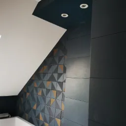 Nowoczesna łazienka z geometryczną mozaiką z płytek w odcieniach szarości i złota, częściowo wykończona, z widoczną białą wanną i oświetleniem punktowym w ciemnym suficie.