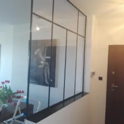 Szklana ścianka działowa w stylu loft z czarnymi szprosami, oddzielająca jadalnię z bukietem czerwonych tulipanów od przedpokoju z ciemnymi drzwiami.