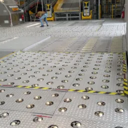 Wnętrze hali magazynowej z podłogą transportową wyposażoną w rolki, widoczne osoby i kontenery DHL w tle.