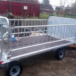 Nowa, ocynkowana przyczepa metalowa z barierkami ochronnymi, stojąca na zewnątrz, z widocznym logo 'WISAG' i czerwoną bramą w tle.