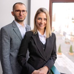 Portret dwojga agentów nieruchomości w eleganckich strojach na tle okna z widokiem na miasto. Kobieta uśmiechnięta, mężczyzna w okularach. Profesjonalizm i zaufanie.