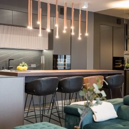 Nowoczesna kuchnia z wyspą i miedzianymi lampami wiszącymi. Minimalistyczny design, ciemne szafki i marmurowy backsplash. Widok z salonu na wysokie krzesła barowe.