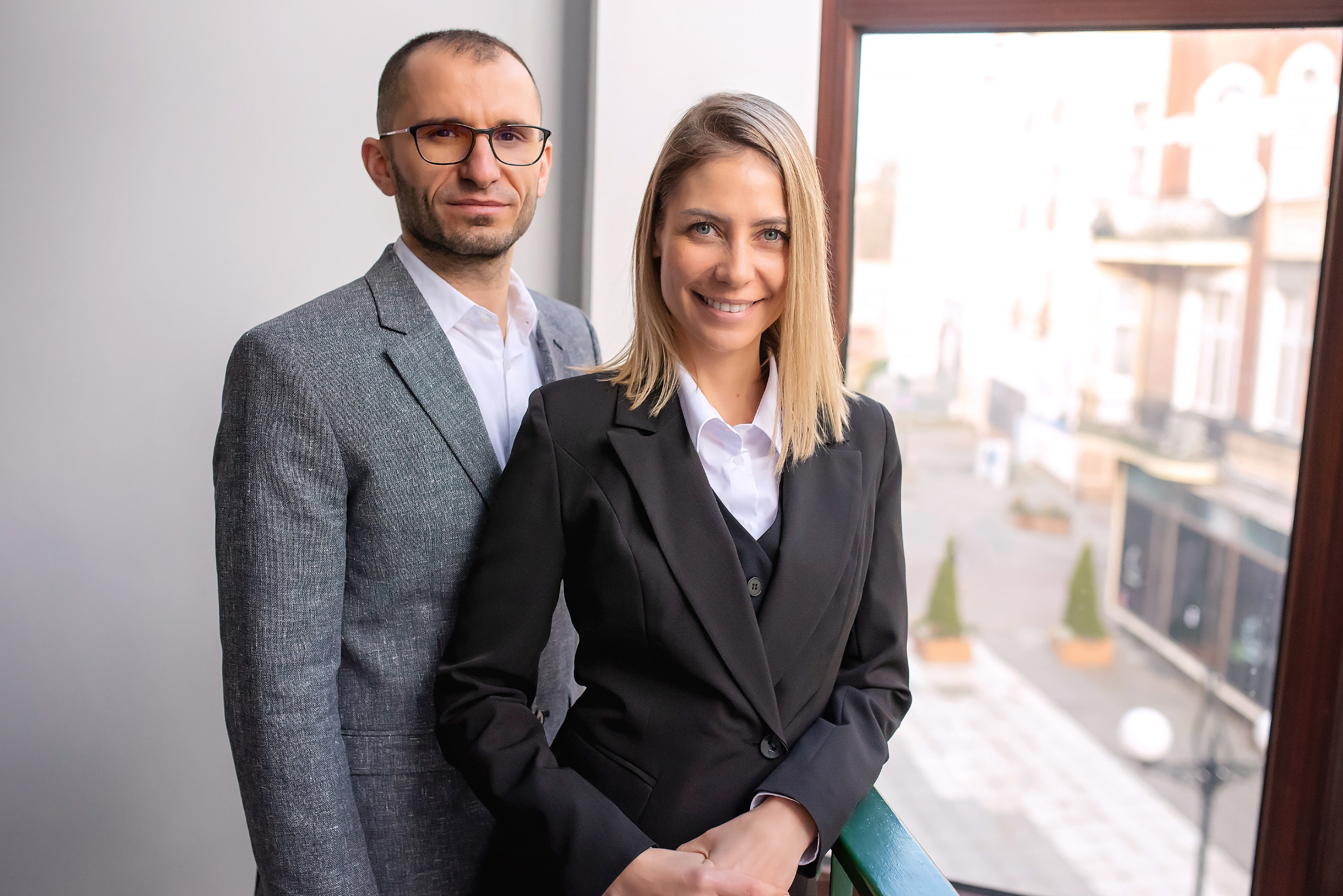 Portret dwojga agentów nieruchomości w eleganckich strojach na tle okna z widokiem na miasto. Kobieta uśmiechnięta, mężczyzna w okularach. Profesjonalizm i zaufanie.