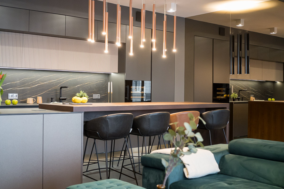 Nowoczesna kuchnia z wyspą i miedzianymi lampami wiszącymi. Minimalistyczny design, ciemne szafki i marmurowy backsplash. Widok z salonu na wysokie krzesła barowe.