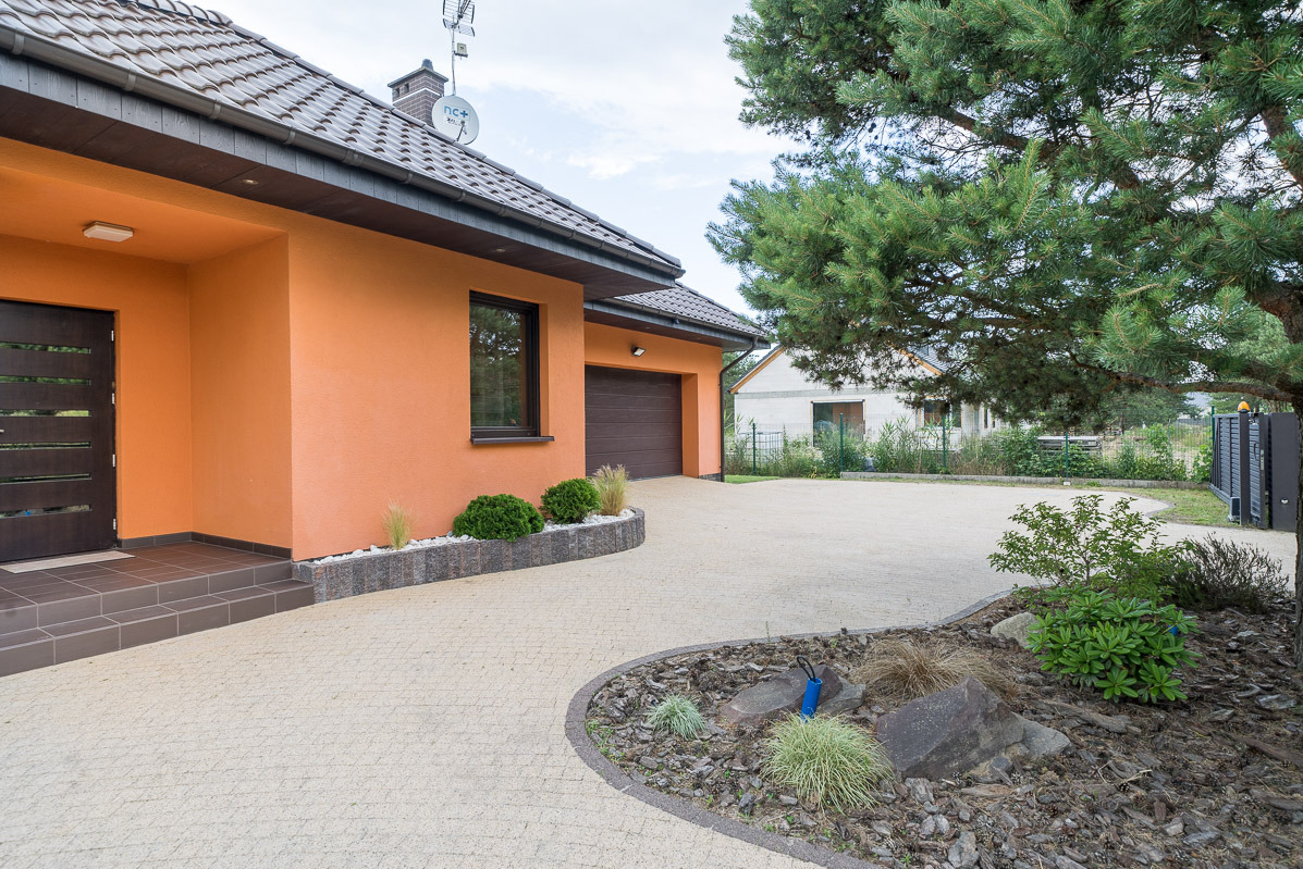 Pomarańczowy dom jednorodzinny z brązowymi akcentami i brukowanym podjazdem w Gorzowie Wielkopolskim, otoczony zielenią. Widok z zewnątrz, w słoneczny dzień.