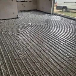 Instalacja ogrzewania podłogowego w surowym stanie, rury ułożone na siatce, widok z góry na dużą powierzchnię z oknem i drzwiami balkonowymi w tle.