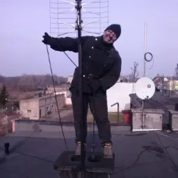 Uśmiechnięty kominiarz w roboczym stroju stoi na kominie obok anteny telewizyjnej, trzymając linkę z obciążnikiem.