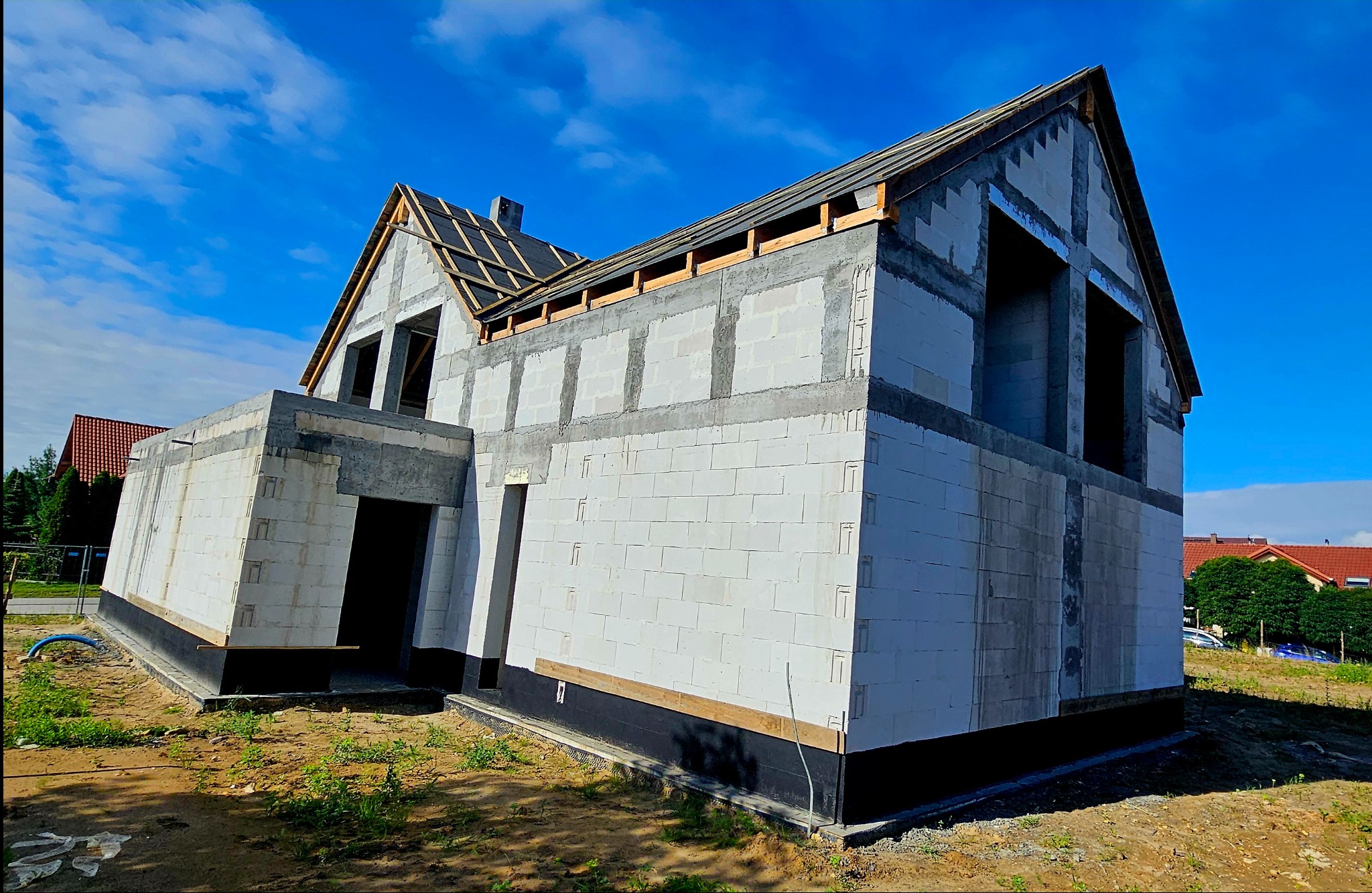 Surowy stan domu z białych bloczków, widoczne elementy konstrukcji dachu, czarne deski na dachu, niebieskie niebo w tle.