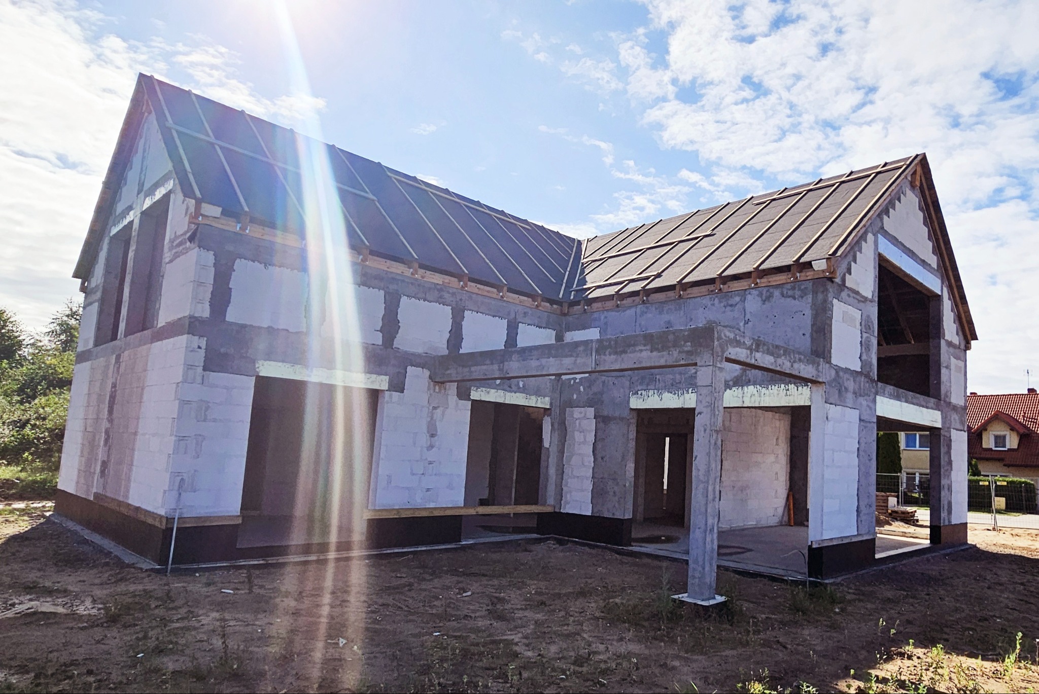 Surowy, dwukondygnacyjny dom murowany w trakcie budowy, z częściowo ukończonym dachem z papy na łatach, widoczne białe bloczki betonowe i betonowe słupy werandy, na działce ziemi.