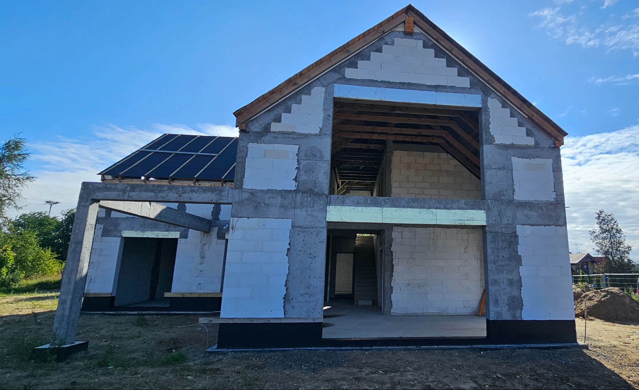 Surowy stan domu z betonu komórkowego z częściowo położonym dachem, widoczne elementy konstrukcyjne oraz otwory okienne i drzwiowe, w tle błękitne niebo.