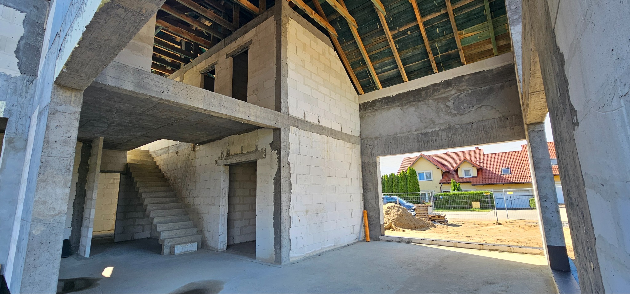 Wnętrze surowego domu w Kolbudach, w trakcie budowy: widoczne ściany z bloczków, betonowe schody prowadzące na piętro, drewniany szkielet dachu, z zewnątrz widok na sąsiednie domy.