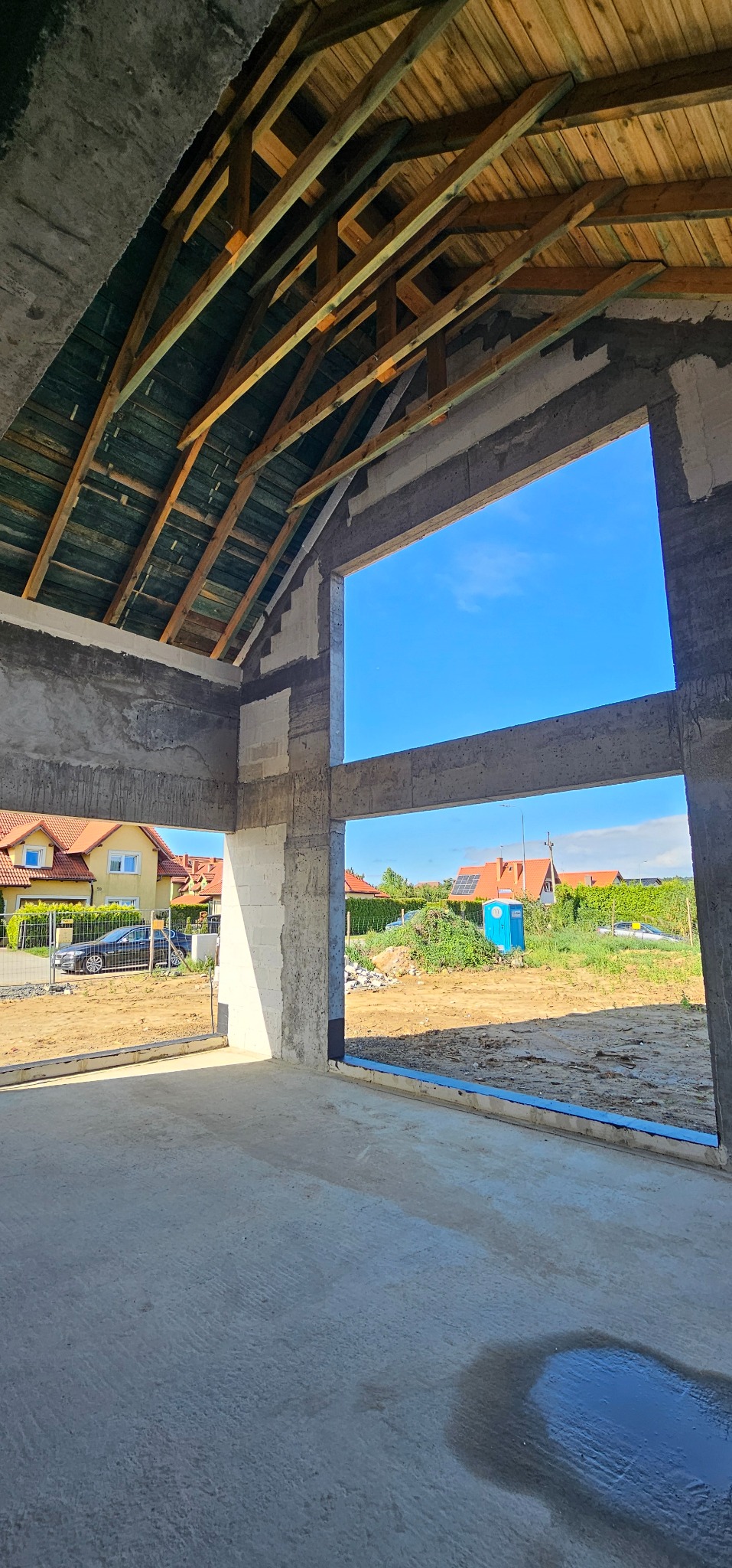 Wnętrze surowego domu w trakcie budowy, widoczne drewniane krokwie dachu, ściany z betonu komórkowego i widok na sąsiednie domy przez otwory okienne.