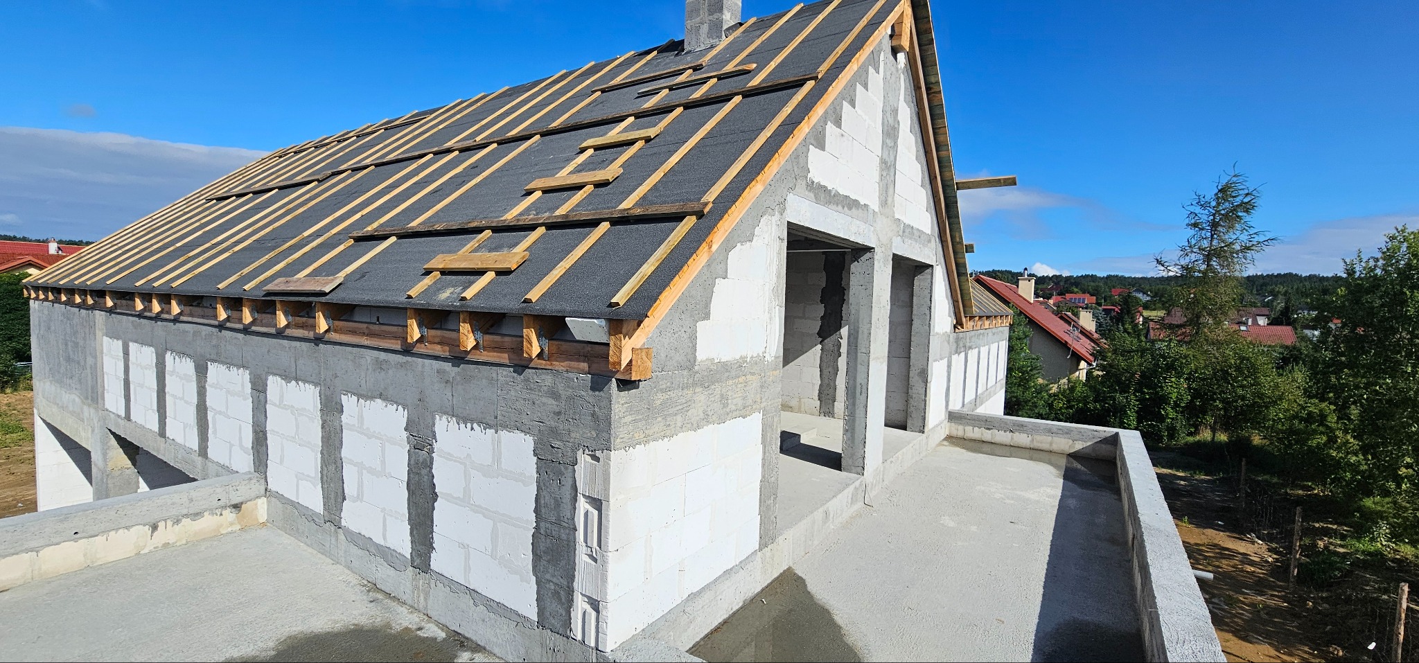 Surowy stan domu z białych bloczków, widoczna konstrukcja dachu z papą i łatami, otwarte przestrzenie okienne i drzwiowe, tło stanowią drzewa i zabudowania.