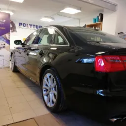 Błyszczący, czarny samochód marki Audi po usłudze detailingowej, widoczny z perspektywy tylnego lewego koła, z odblaskami światła na lakierze i banerami reklamowymi w tle.