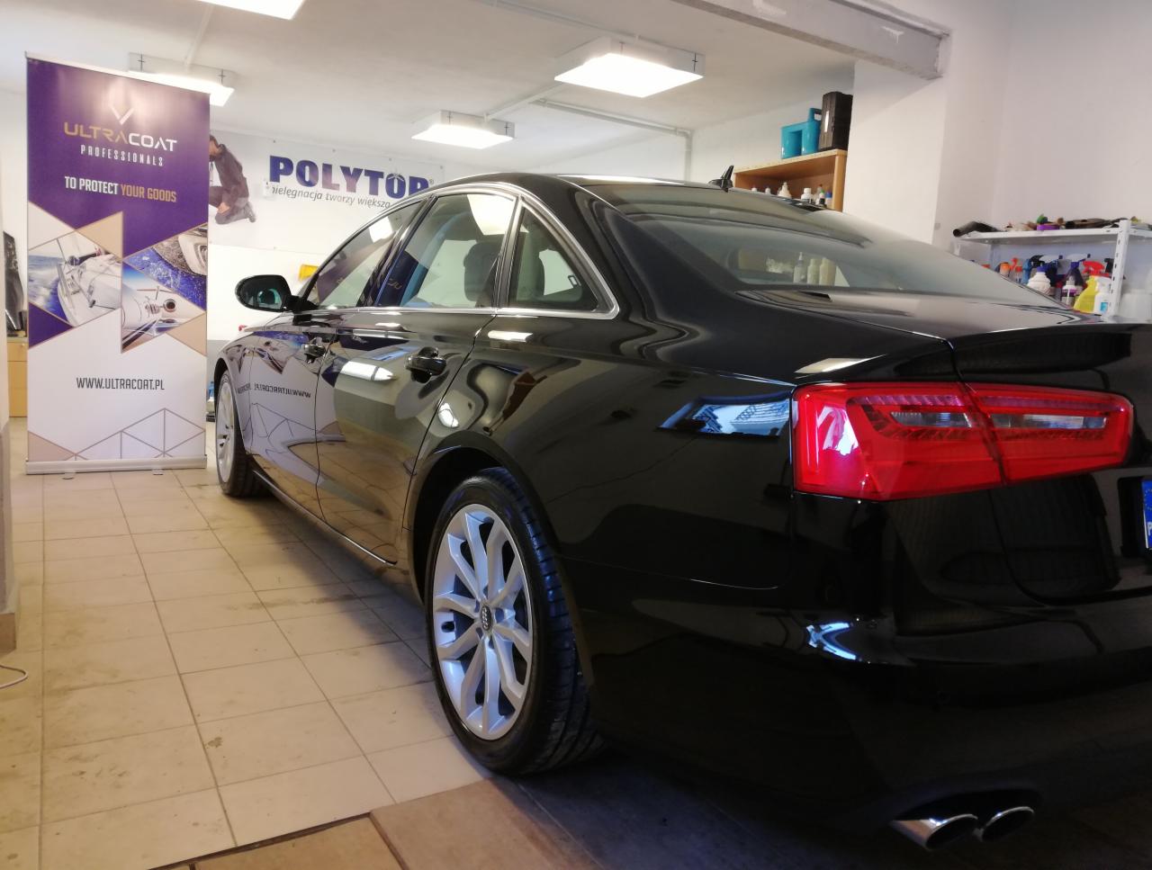 Błyszczący, czarny samochód marki Audi po usłudze detailingowej, widoczny z perspektywy tylnego lewego koła, z odblaskami światła na lakierze i banerami reklamowymi w tle.