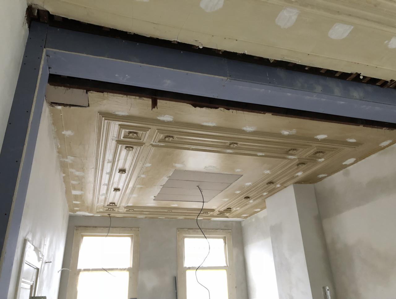 Pokój w trakcie prac remontowych: dekoracyjny sufit z gipsowymi zdobieniami, częściowo pokryty szpachlą, widoczna stalowa belka wzmacniająca konstrukcję, ściany przygotowane do malowania, dwa okna...