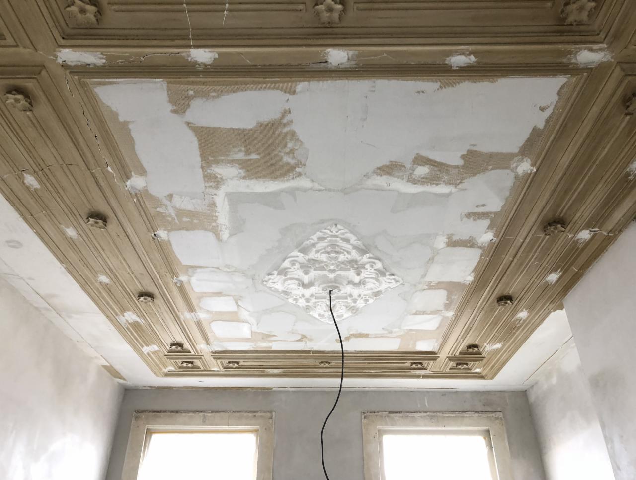 Sufit w trakcie renowacji z dekoracyjnymi elementami sztukaterii i widocznymi śladami po szpachlowaniu, podkreślony przez naturalne światło wpadające przez okno.