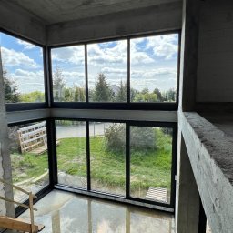 Locuss (Okna Oknoplast Warszawa) - Dwukondygnacyjne okno aluminiowe w surowym stanie budynku, widok z góry na drewniane schody i podłogę z kałużami. Za oknem zieleń i błękitne niebo.