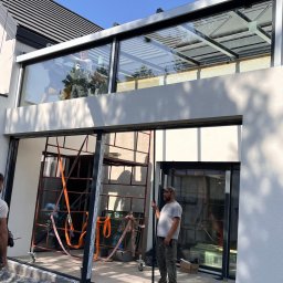 Locuss (Okna Oknoplast Warszawa) - Montaż panoramicznych okien przesuwnych w nowoczesnym domu. Widoczni dwaj monterzy i rusztowanie. Duże przeszklenia z widokiem na zewnątrz.