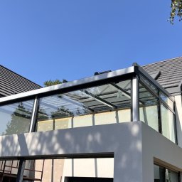 Locuss (Okna Oknoplast Warszawa) - Nowoczesna zabudowa tarasu z aluminiową konstrukcją i szklanymi panelami, widok z dołu, z elementami rusztowania i dźwigu w tle, błękitne niebo.
