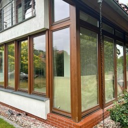 Locuss (Okna Oknoplast Warszawa) - Zewnętrzny widok przeszklonej werandy z drewnianą ramą okienną, z widoczną architekturą domu i ogrodem w tle. Ujęcie z poziomu gruntu, podkreślające detale wykończenia.