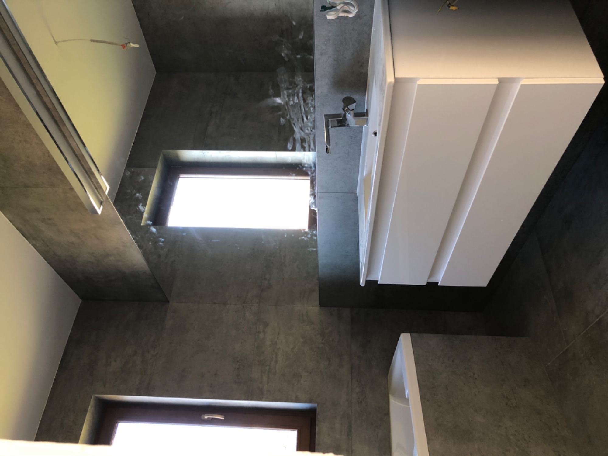 Szara łazienka z oknem, białą szafką pod umywalką i baterią. Widoczne płytki imitujące beton na ścianach i podłodze. Niezakończone prace elektryczne na suficie.