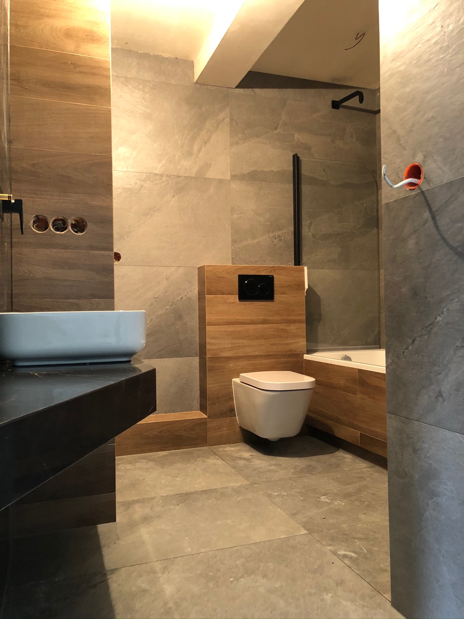 Nowoczesna łazienka z płytkami imitującymi drewno i kamień, wisząca toaleta z czarnym przyciskiem, umywalka nablatowa na czarnym blacie oraz widoczne otwory pod baterię.