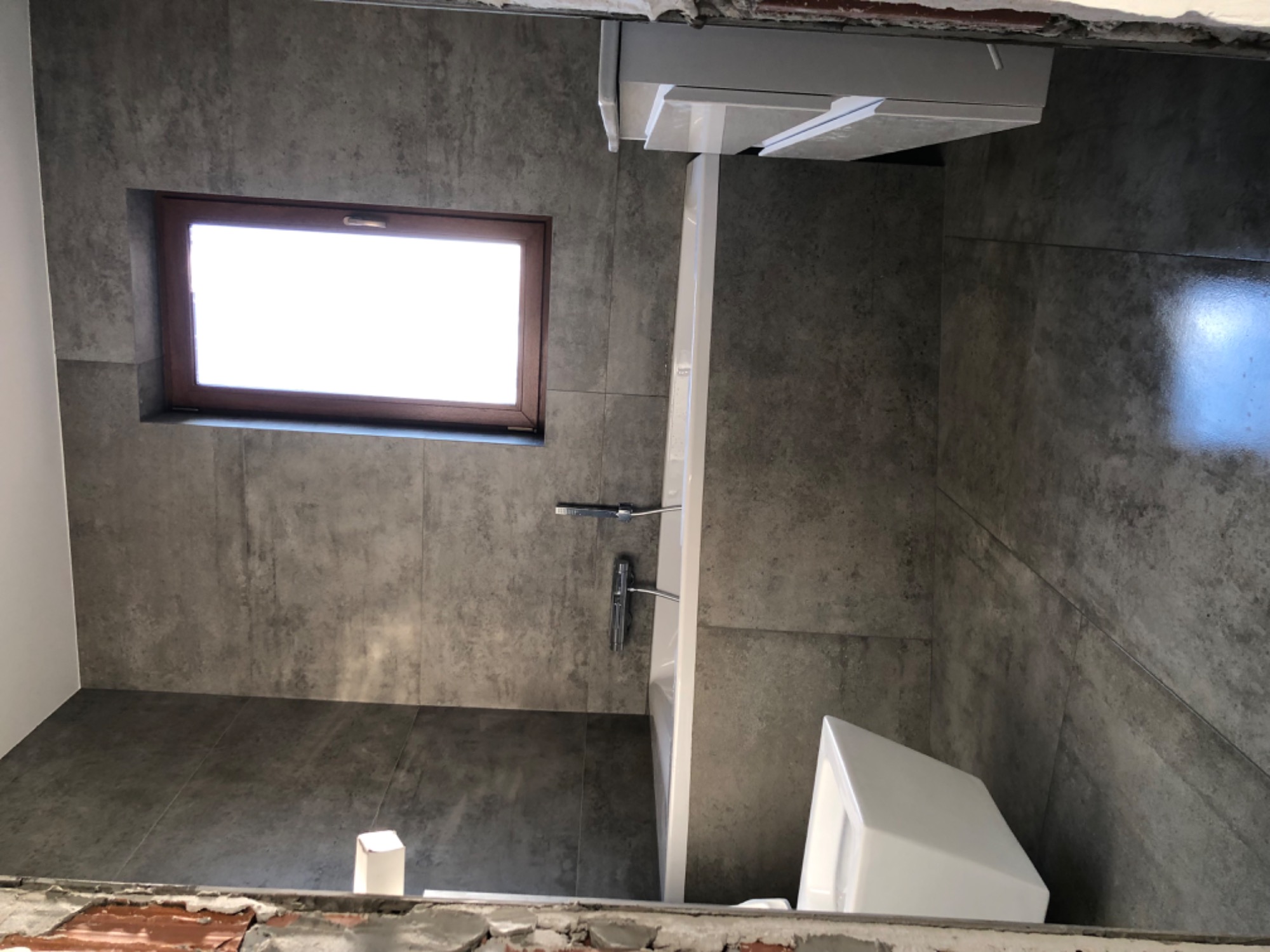 Łazienka w trakcie remontu z szarymi płytkami imitującymi beton, widoczne okno z drewnianą ramą, biała umywalka i częściowo zamontowana armatura.