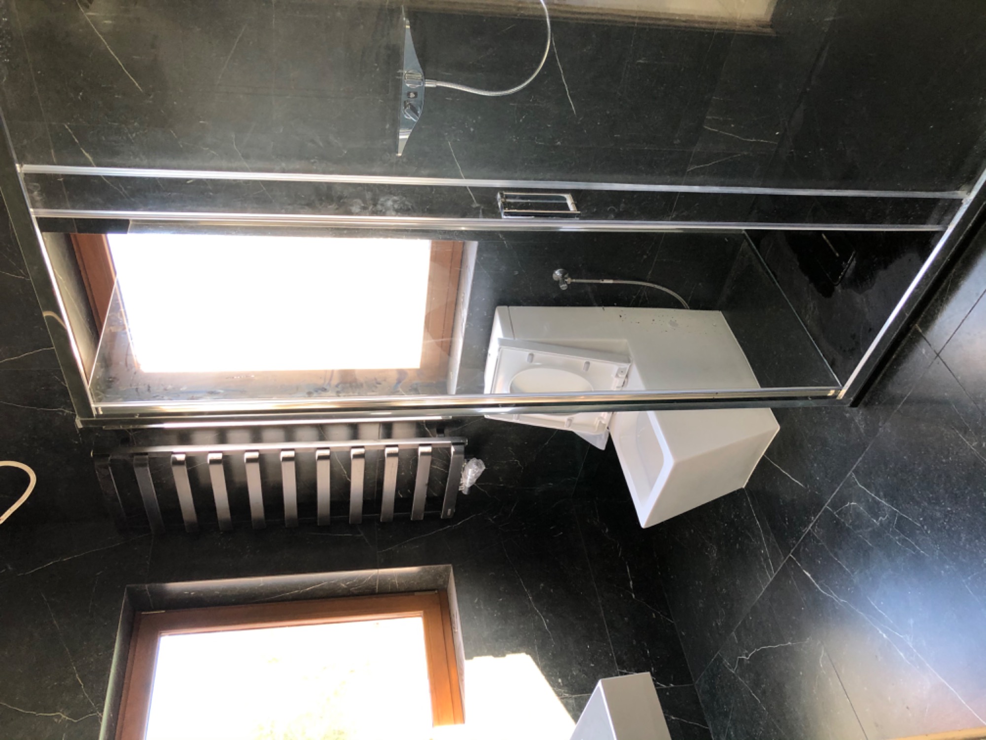 Ciemna, marmurkowa łazienka z kabiną prysznicową, umywalką i grzejnikiem drabinkowym pod oknem. Widoczne białe elementy armatury na tle ciemnych płytek. Na podłodze leży folia.