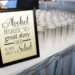 Czarna ramka z napisem 'Alcohol because no great story ever began with a salad' na stole z rzędem pustych szklanek, przygotowanych do serwowania napoj&oacute;w.