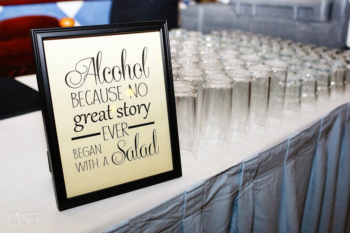 Czarna ramka z napisem 'Alcohol because no great story ever began with a salad' na stole z rzędem pustych szklanek, przygotowanych do serwowania napojów.