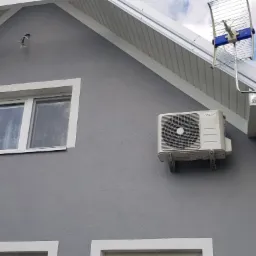 Szara ściana domu z zamontowaną jednostką zewnętrzną klimatyzacji MDV, biała stolarka okienna i antena telewizyjna na dachu.