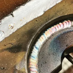 Zbliżenie na świeżo wykonany, wielobarwny spaw na metalowej konstrukcji, ukazujący strukturę i przebarwienia termiczne.