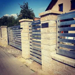 Nowoczesna brama wjazdowa z poziomych, metalowych profili, osadzona na kamiennych słupkach i murku, z widoczną skrzynką na listy, na tle domu jednorodzinnego.