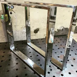 Dwie, lustrzanie wypolerowane, prostokątne ramy z metalu, prawdopodobnie ze stali nierdzewnej, ustawione na perforowanym stole spawalniczym, z widocznym butlą z gazem i reduktorem w tle.