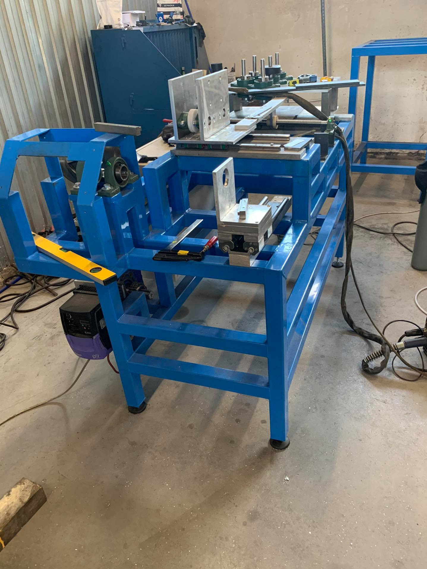 Niebieska, metalowa konstrukcja urządzenia warsztatowego z elementami do obróbki metalu, w tle widoczny sprzęt warsztatowy i narzędzia.