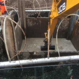 Głęboki wykop zabezpieczony stalowymi szalunkami, wypełniony wodą; żółty wysięgnik koparki JCB zagłębia łyżkę w błotnistej cieczy; na powierzchni ziemi widoczne rury i pracownicy w odblaskowych...